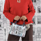 A person wearing a bright red knit sweater over a plaid collared shirt and dark brown pants, holding a small illustrated handbag in front of a black-and-white architectural cityscape backdrop.
