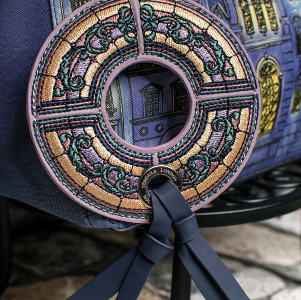 Close-up of a handbag featuring an intricate circular embroidered design in gold, purple, and green, with a navy blue leather strap and a matching printed architectural pattern on the bag body.