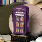 A purple telephone-shaped book swap box styled like a Tinted Telephone Booth, decorated with miniature book illustrations on its front window panels. The box is displayed on a stack of vintage-style books in a cozy, styled home environment with pillows and open books in the background.
