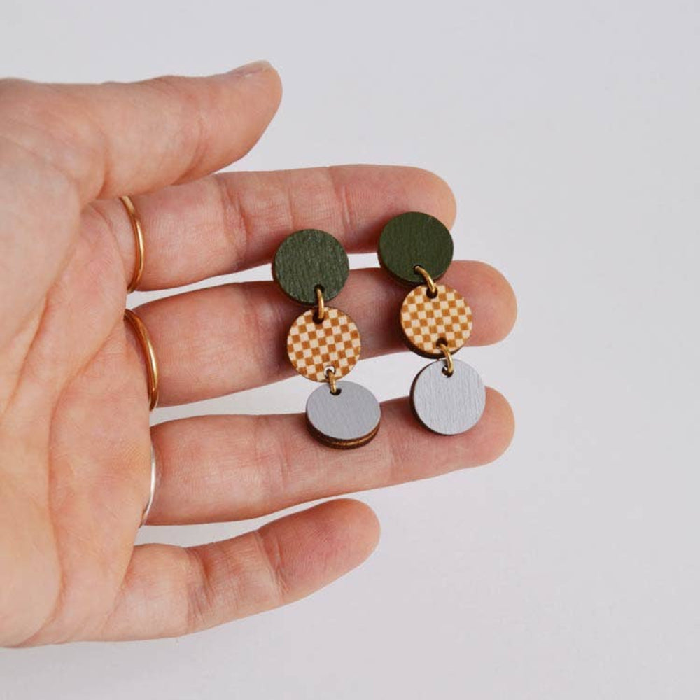 Hand holding a pair of earrings with green, gold, and silver circular components on a light background