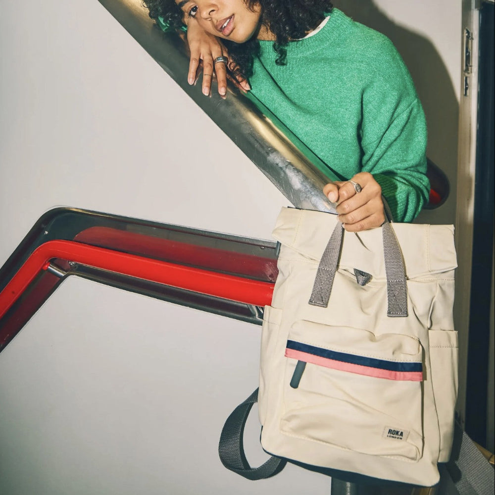 A person in a green sweater holds a beige tote bag with gray handles and a pink and navy stripe detail.