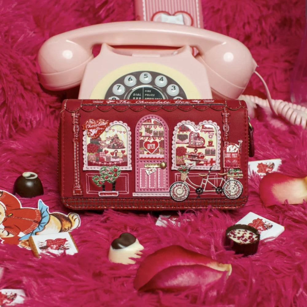 Red vintage-style wallet with intricate designs on a pink textured background with a pink rotary phone and candy.