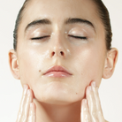 Close-up of a person with closed eyes and hands touching their face, showing a glossy, hydrated skincare finish against a plain light background.