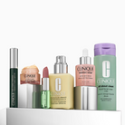 Six Clinique skincare products arranged on a white pedestal against a light gray background, including a dark green eye product, a peach-colored cream, a pink tube, a yellow moisturizer bottle, a silver-toned serum bottle, and a mint green body wash bottle.