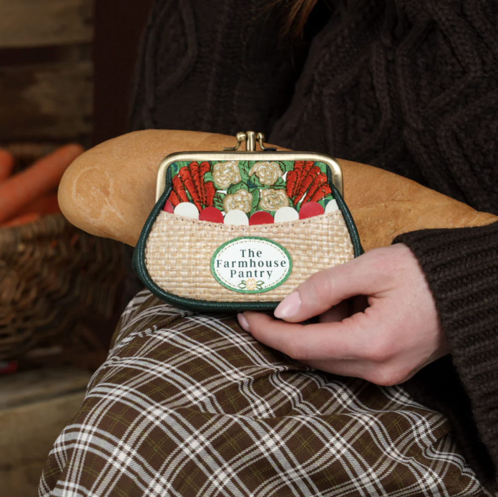 A person holds a small woven purse shaped like a basket of vegetables, with a visible label reading "The Farmhouse Pantry." The scene includes the person's hand, torso, and plaid skirt in a real indoor setting, so this is Lifestyle rather than Product/Packaging.