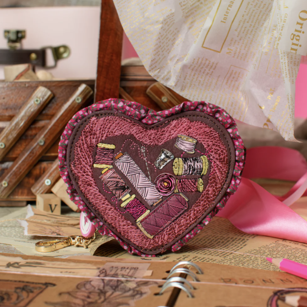 A heart-shaped fabric coin purse with a ruffled edge, featuring an applique design of sewing spools of thread in shades of pink, purple, and brown. The purse is styled on a desk with vintage books, a pink ribbon, and a notebook.
