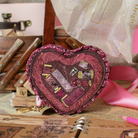 A heart-shaped fabric coin purse with a ruffled edge, featuring an applique design of sewing spools of thread in shades of pink, purple, and brown. The purse is styled on a desk with vintage books, a pink ribbon, and a notebook.