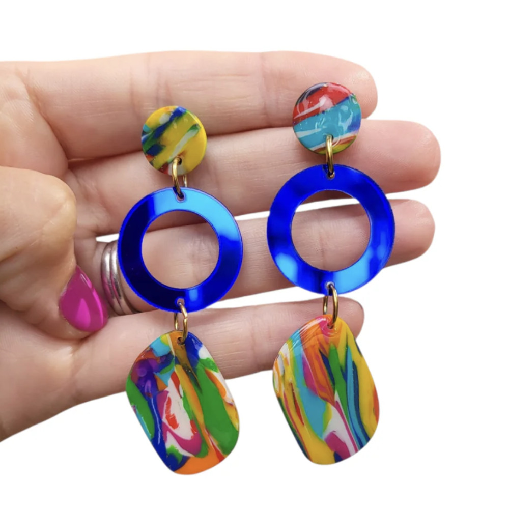 Colorful earrings with blue circular middle section held in a hand against a white background
