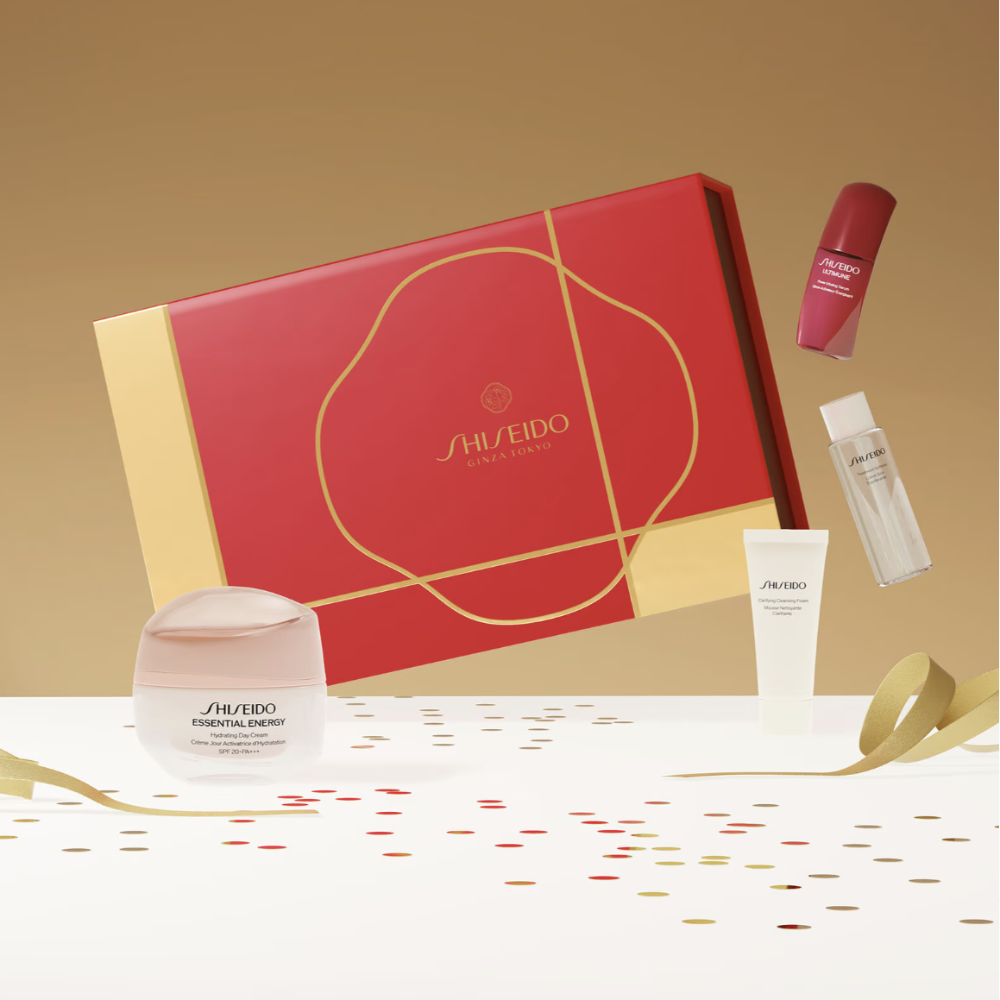 Shiseido gift set featuring a red and gold box surrounded by skincare products including a jar, tube, and bottle on a white surface with gold ribbons and confetti, against a warm beige background.