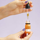 Hand holding a dropper with face oil over a bottle of face oil against a purple background