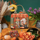 A decorative handbag with an illustrated autumn storefront scene, styled in a fall-themed arrangement with pumpkins, flowers, and draped orange fabric against a dark green background.