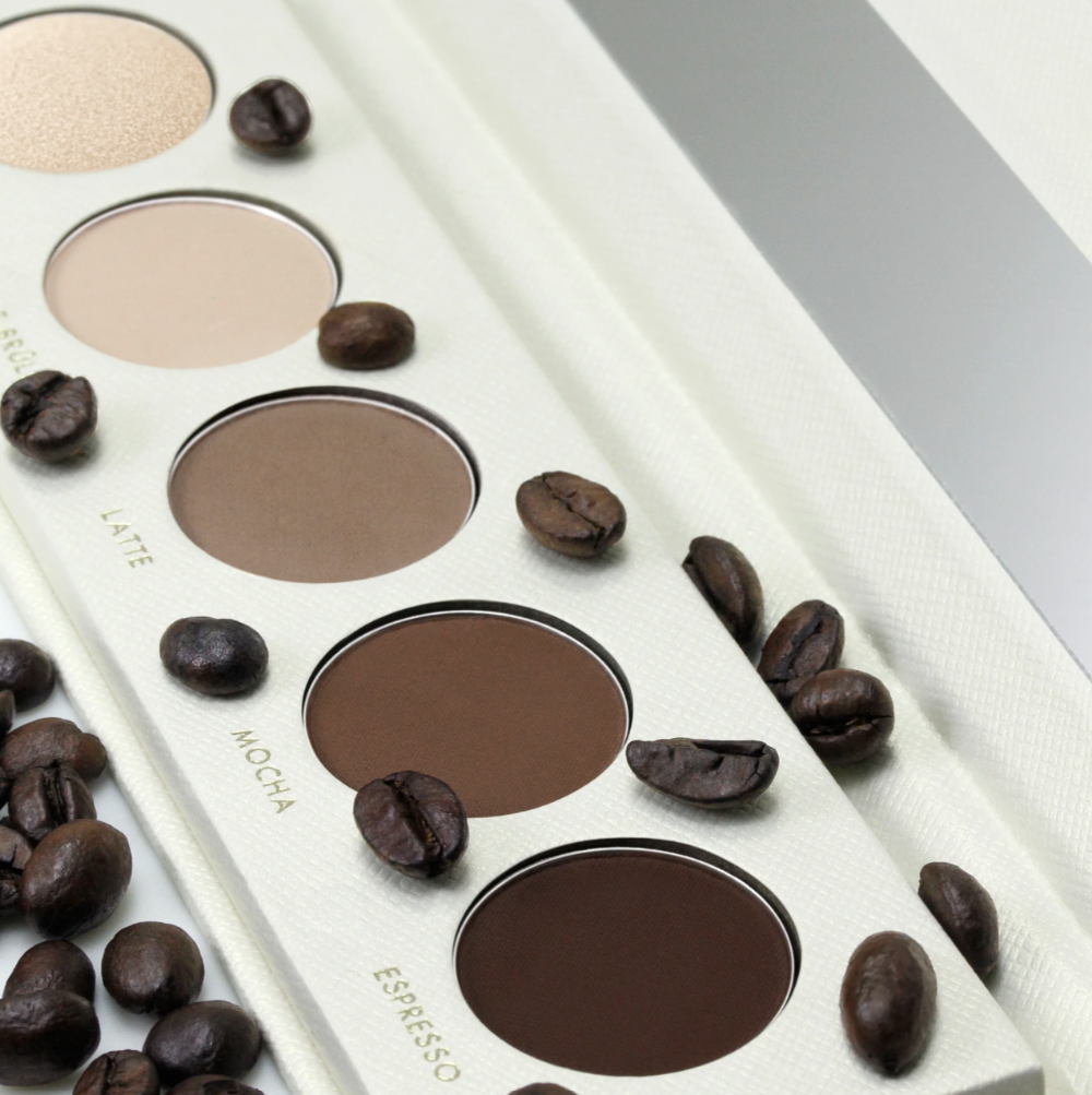 A close-up of a makeup eyeshadow palette with shades labeled Latte, Mocha, and Espresso, styled with scattered coffee beans on a white textured background.