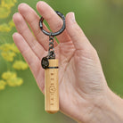 Hand holding a wooden keychain with 'BEEVIVE' branding against a blurred natural background.