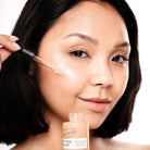 Woman applying a cosmetic product to her face with a brush, holding a bottle labeled 'Pimele'.