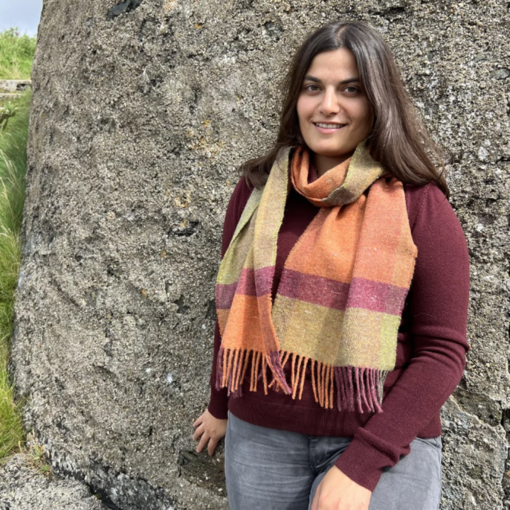A woman wearing a maroon long-sleeve top and gray jeans stands outdoors against a large textured rock, wearing a thick woven scarf with orange, yellow, and pink stripes and fringe.