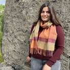A woman wearing a maroon long-sleeve top and gray jeans stands outdoors against a large textured rock, wearing a thick woven scarf with orange, yellow, and pink stripes and fringe.
