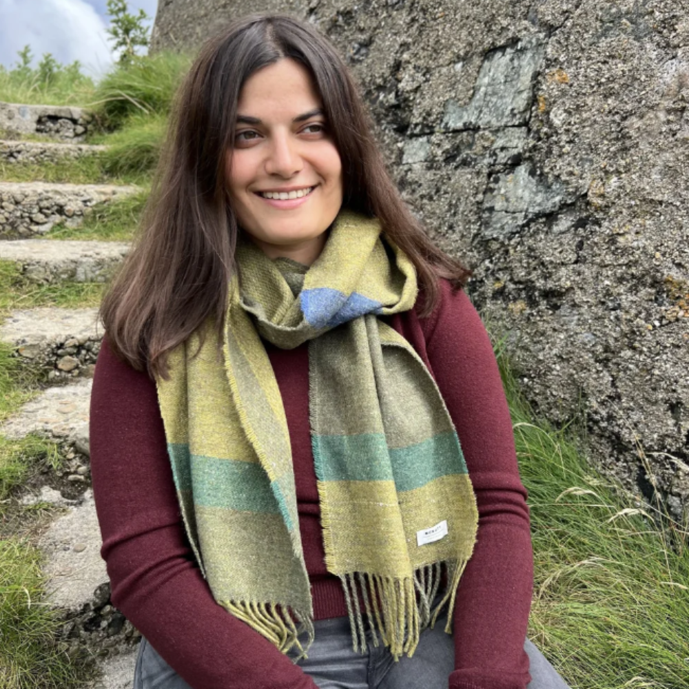 A woman sitting outdoors in front of a stone wall, wearing a multicolor striped knit scarf with green, yellow, and blue tones over a burgundy top and gray pants.
