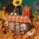 A decorative handbag styled like a storefront with autumn-themed printed panels, a scalloped orange roof, floral trim, and a dark handle, surrounded by pumpkins, flowers, and draped gold fabric against a teal background.