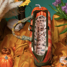 A decorative handbag styled in an autumn scene with pumpkins, flowers, and draped gold fabric. The bag has a pumpkin-shaped top, orange and white stripes on the exterior, a green handle with sunflower details, and a patterned interior.