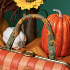 A close-up of a handbag handle in muted olive green with embroidered sunflowers, set against a styled autumn scene with pumpkins, a yellow flower, and orange fabric.