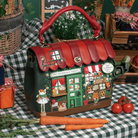 A decorative handbag shaped like a small shop front, with a red handle and a multicoloured illustrated facade, displayed on a green-and-white checkered tablecloth with carrots, tomatoes, baskets, and plants around it.