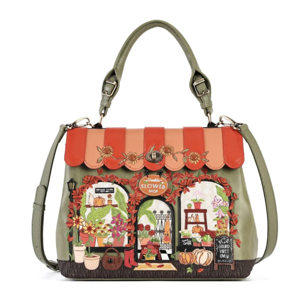 A structured handbag with olive green leather trim and handles, featuring a large, colorful embroidered design of a flower shop on the front. The bag has a scalloped orange and peach embroidered awning with floral details, and a multicolor storefront scene below.
