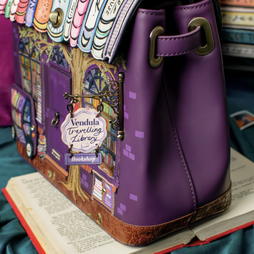 A purple handbag with a whimsical library-themed design, featuring book spines along the top, a detailed illustration of a library facade, and a hanging sign that reads 'Vendula travelling library bookshop.' The bag is styled on top of an open book against a dark teal fabric background.