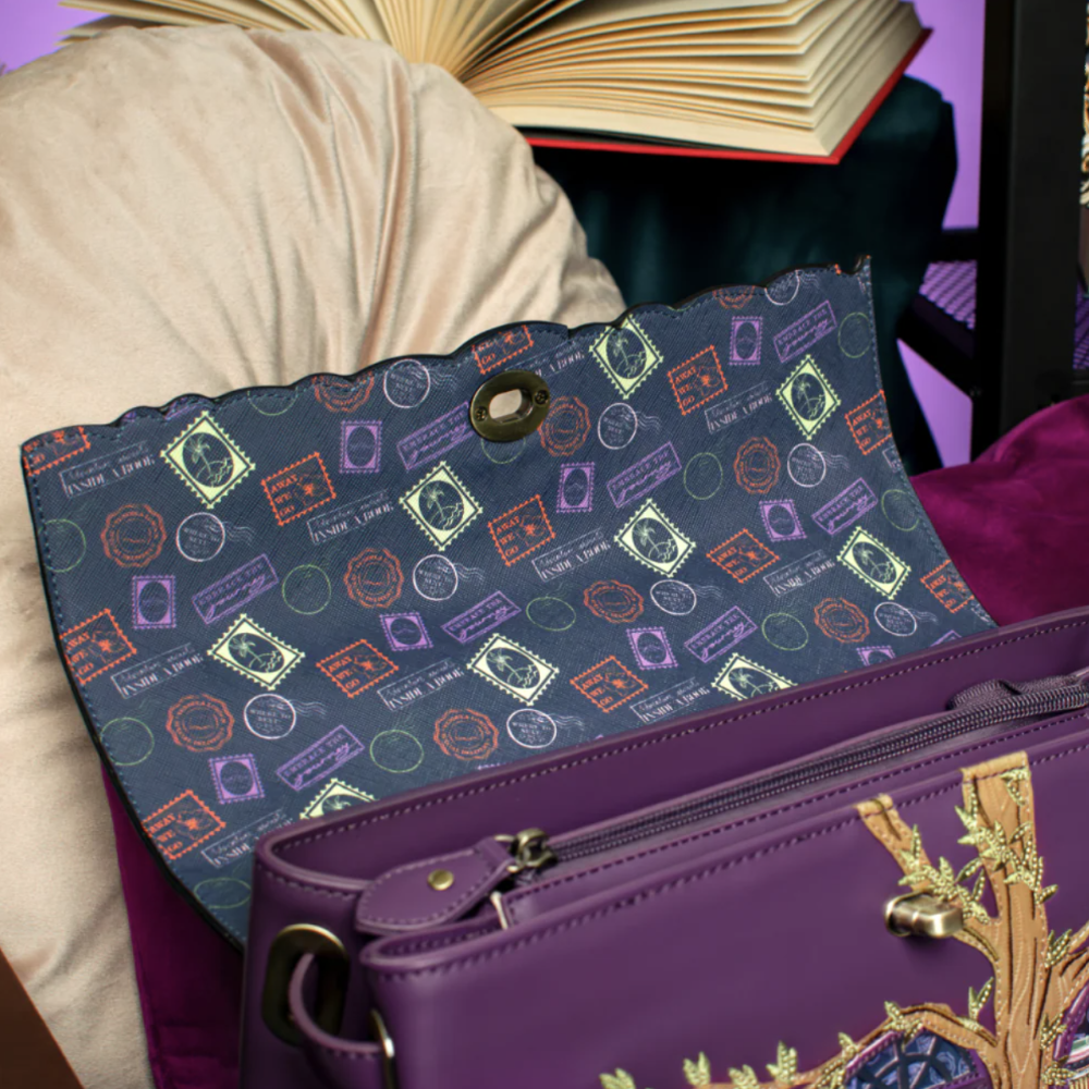 A navy blue fabric pouch with a colorful stamp pattern and a gold snap button, resting on a purple leather handbag with gold embroidery in a styled setting.