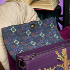 A navy blue fabric pouch with a colorful stamp pattern and a gold snap button, resting on a purple leather handbag with gold embroidery in a styled setting.
