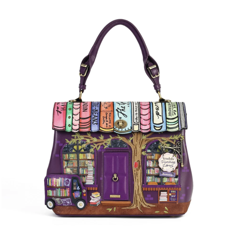 A purple handbag designed to look like a whimsical bookshop, featuring colorful book spines across the top, a purple door, a tree motif, and a small book truck on the side, shown on a plain white background.