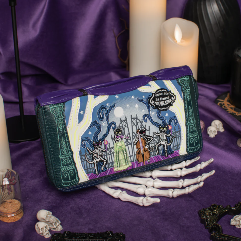 A purple zip-around wallet with a detailed gothic skeleton illustration, styled on a purple fabric surface with candles, skeleton props, and skulls.
