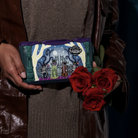 A person in a brown leather jacket and brown knit top holds a small illustrated zip wallet featuring a fantasy scene. The person also holds three red roses against a dark background.
