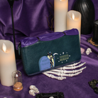 A dark teal and purple faux-leather wallet with a Halloween-themed cat-and-tombstone graphic, styled on a purple fabric background with candles, skeleton bones, and occult props.