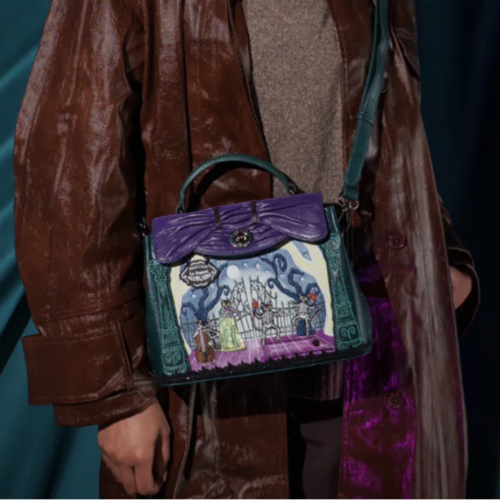 A person wearing a brown leather coat carries a structured handbag with a purple flap, dark teal body, and a colorful illustrated front panel against a dark teal draped backdrop.