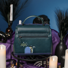 A structured teal leather handbag with a top handle, front zip pocket, and a small gothic tombstone charm, displayed on a black stand with candles, purple fabric, and dark decorative props against a blue background.