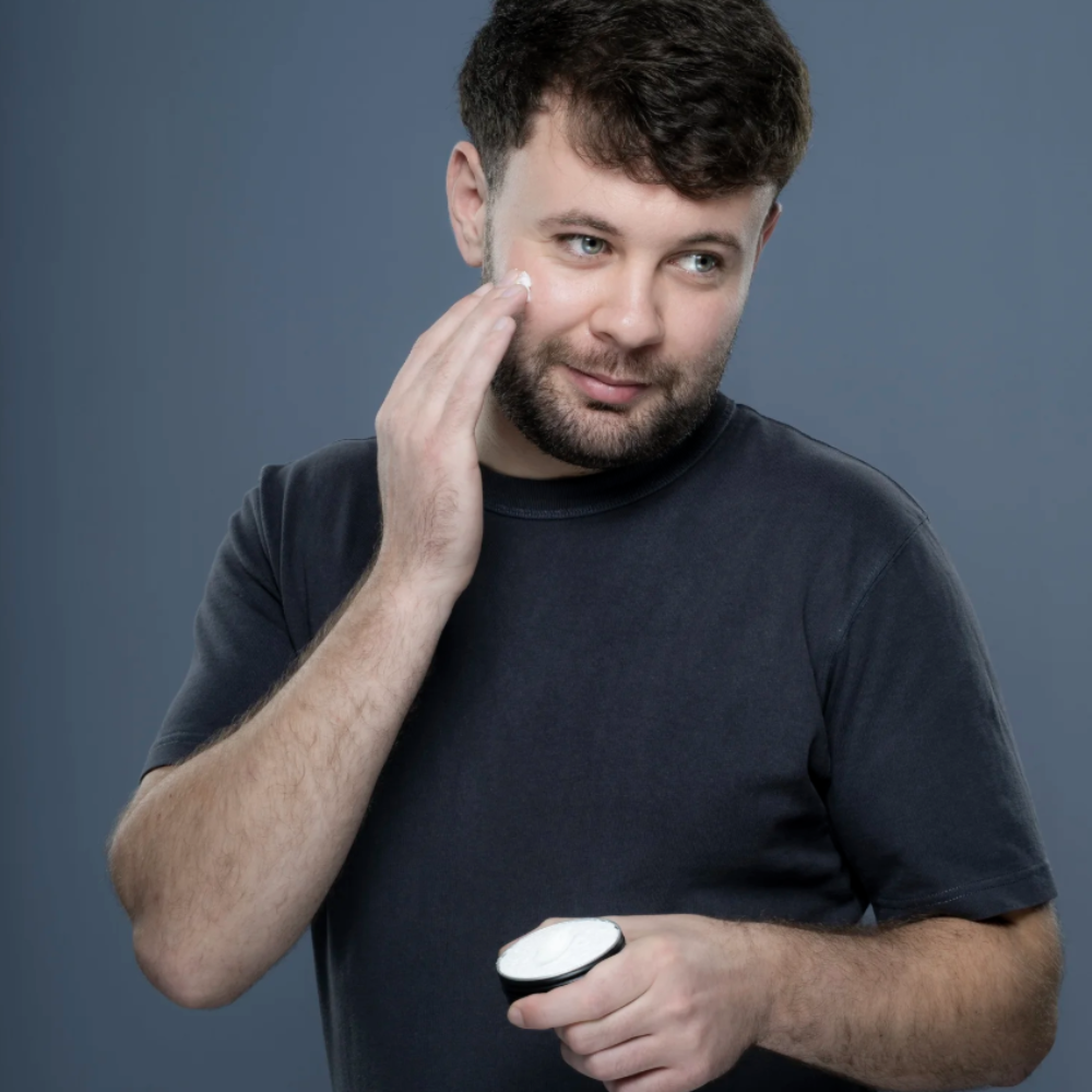A man with short brown hair and a beard applies a white cream from a small black-and-white container to his cheek, wearing a dark gray t-shirt against a solid slate gray background.