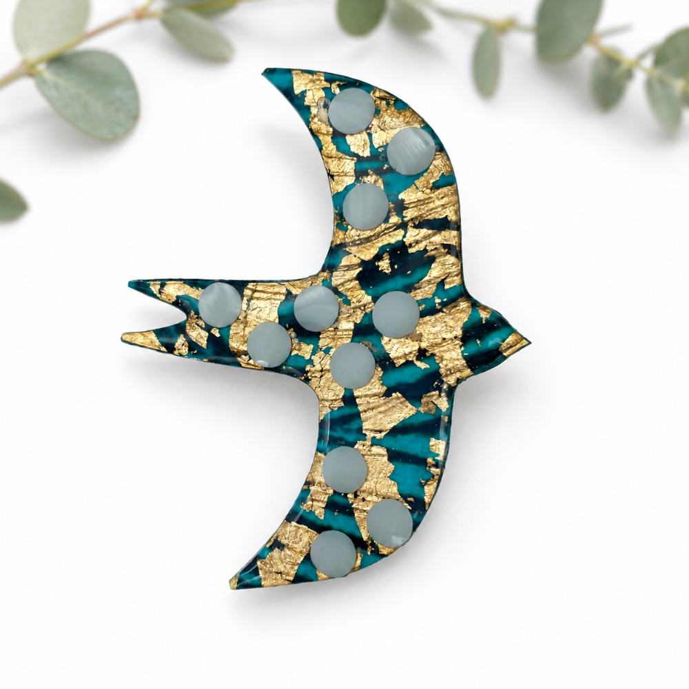 A swallow-shaped brooch with a teal and gold leaf pattern and gray circular studs, photographed on a white background with blurred green leaves in the corner.