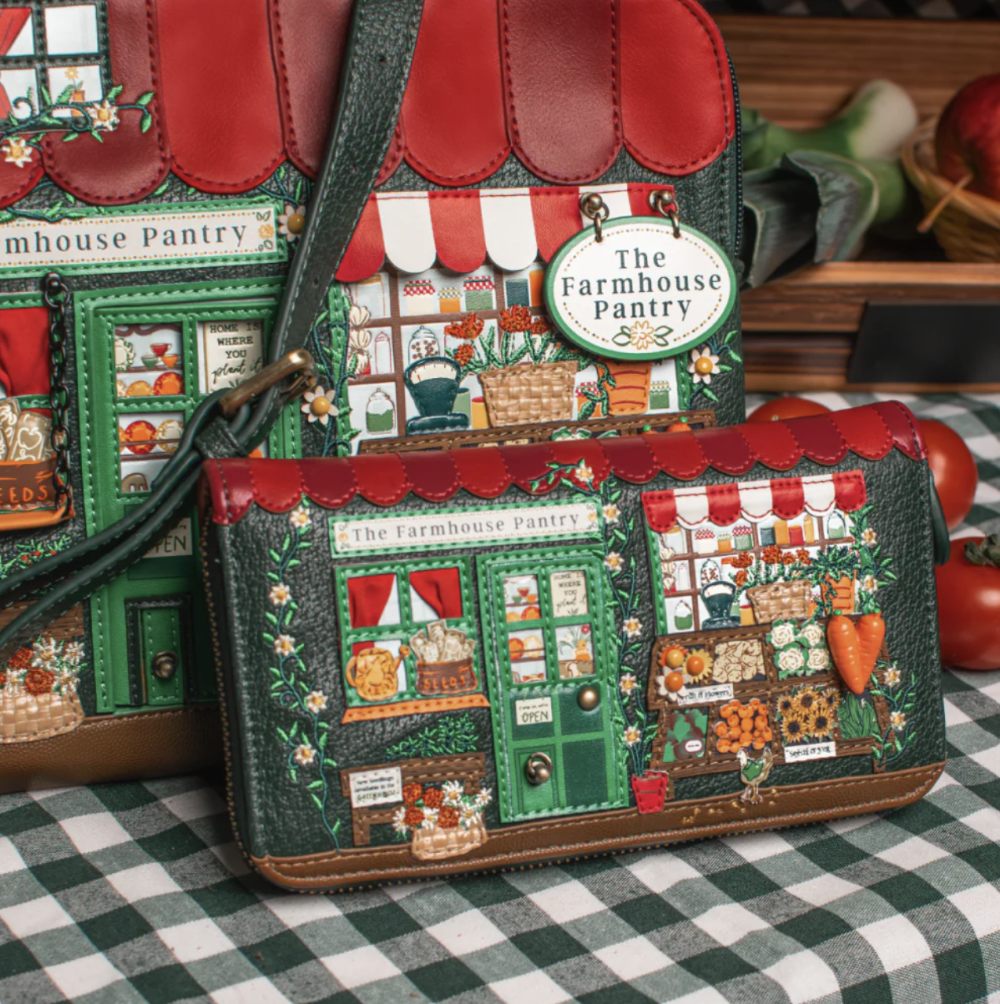 A small crossbody bag with a printed storefront design reading 'The Farmhouse Pantry' sits on a checkered tablecloth. A matching wallet is placed in front, and part of a larger matching bag is visible behind it.