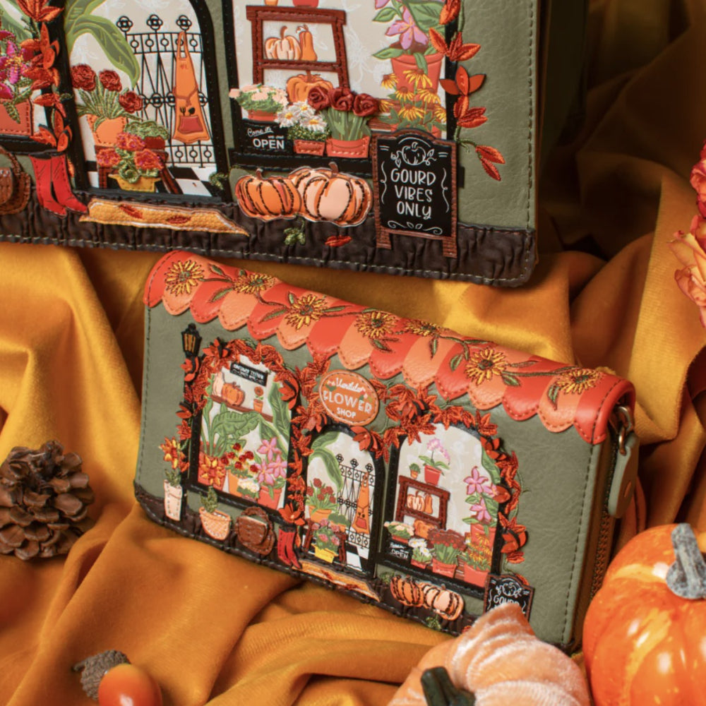 Two olive green faux-leather wallets with autumn-themed storefront embroidery, styled on draped mustard yellow fabric with pumpkins and a pine cone.