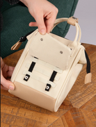 A cream-colored mini tote bag with a pebbled texture, held by hands over a wooden table. The bag features a top handle, a front flap pocket with silver rivets, and black-and-white elastic strap details on the front.