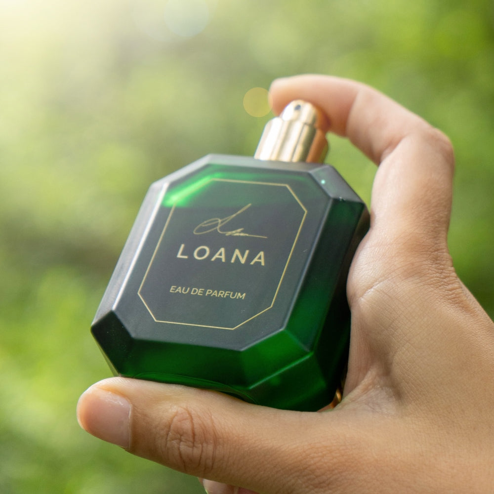 Hand holding a green LOANA perfume bottle against a blurred natural background