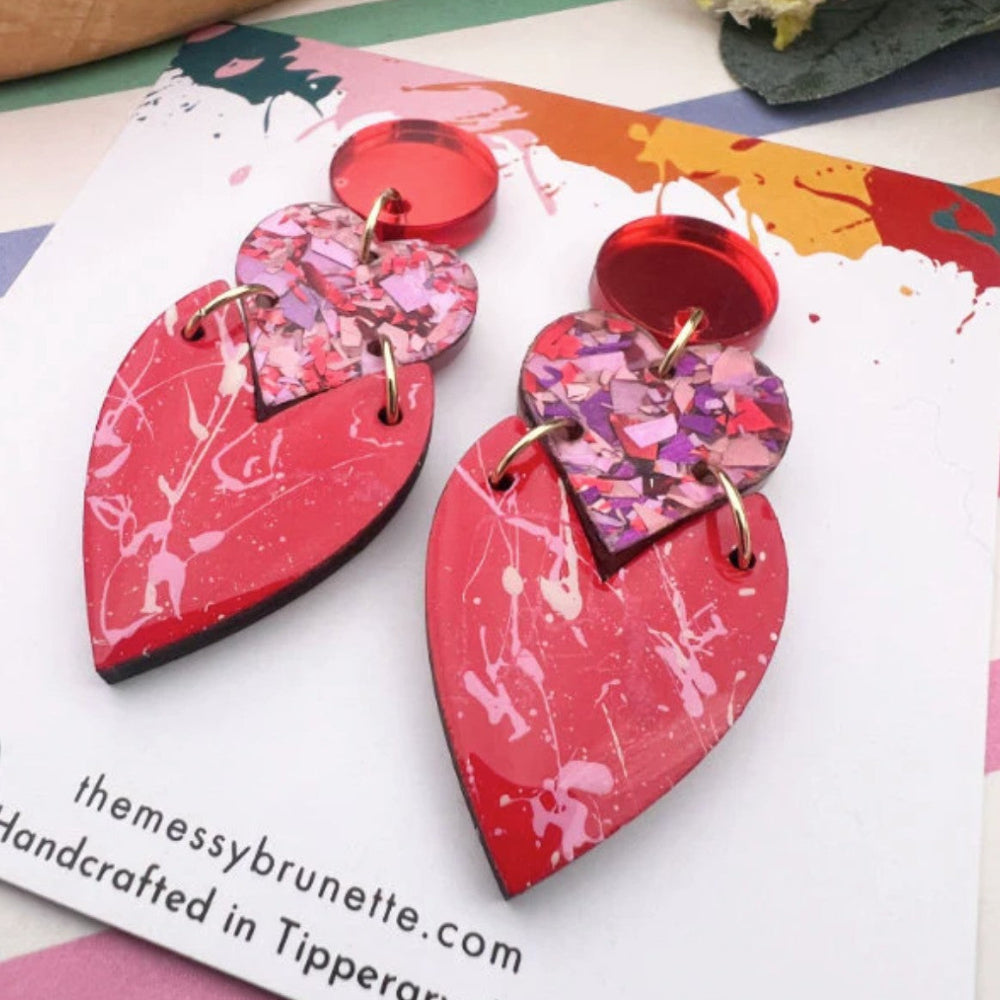Heart-shaped earrings with red and pink marbled design on a colorful card with branding text.