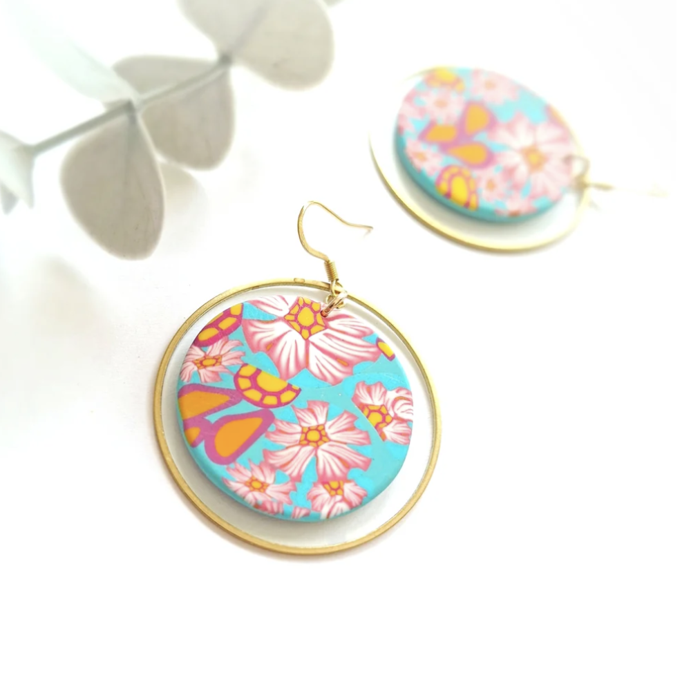 Gold-tone hoop earrings with circular turquoise blue inserts featuring a retro pink, orange, and yellow floral pattern, displayed on a clean white surface with soft leaf shadows.