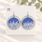 A pair of silver-toned hoop earrings with circular blue and white leaf-patterned inserts, displayed on a white card labeled 'IRISH MADE' against a softly lit neutral background with blurred greenery.