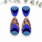 A pair of geometric drop earrings featuring a deep blue top section and a larger teardrop-shaped lower section with a glossy blue gradient and a textured metallic copper and black mosaic center, displayed on a clean white background with soft eucalyptus leaves in the background.