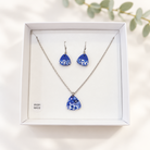 A jewellery set featuring a silver-tone necklace and matching drop earrings with blue and white floral patterned teardrop pendants, presented in a white gift box with 'IRISH MADE' printed on the inside.