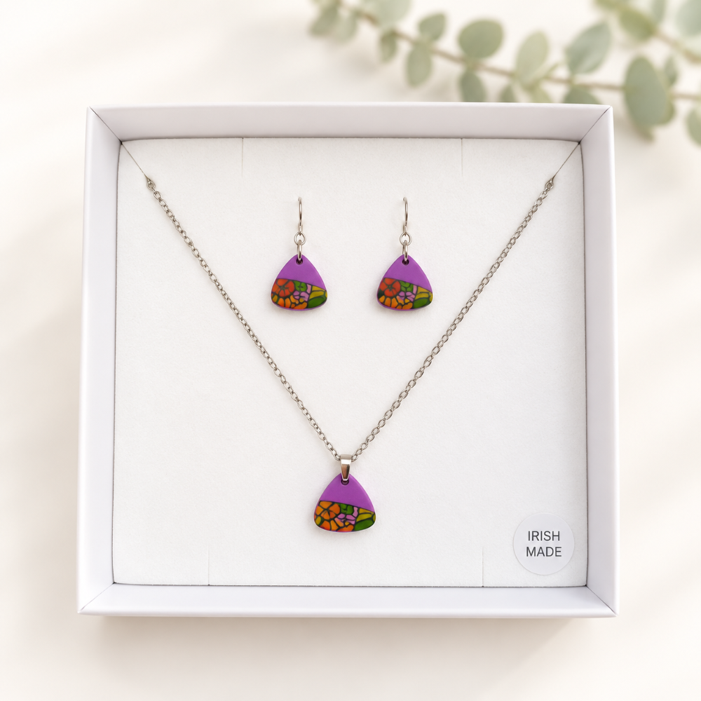 A jewelry set in a white gift box featuring a silver necklace and matching drop earrings with purple teardrop pendants decorated with colorful floral patterns. The box has a small 'IRISH MADE' label on the bottom right.