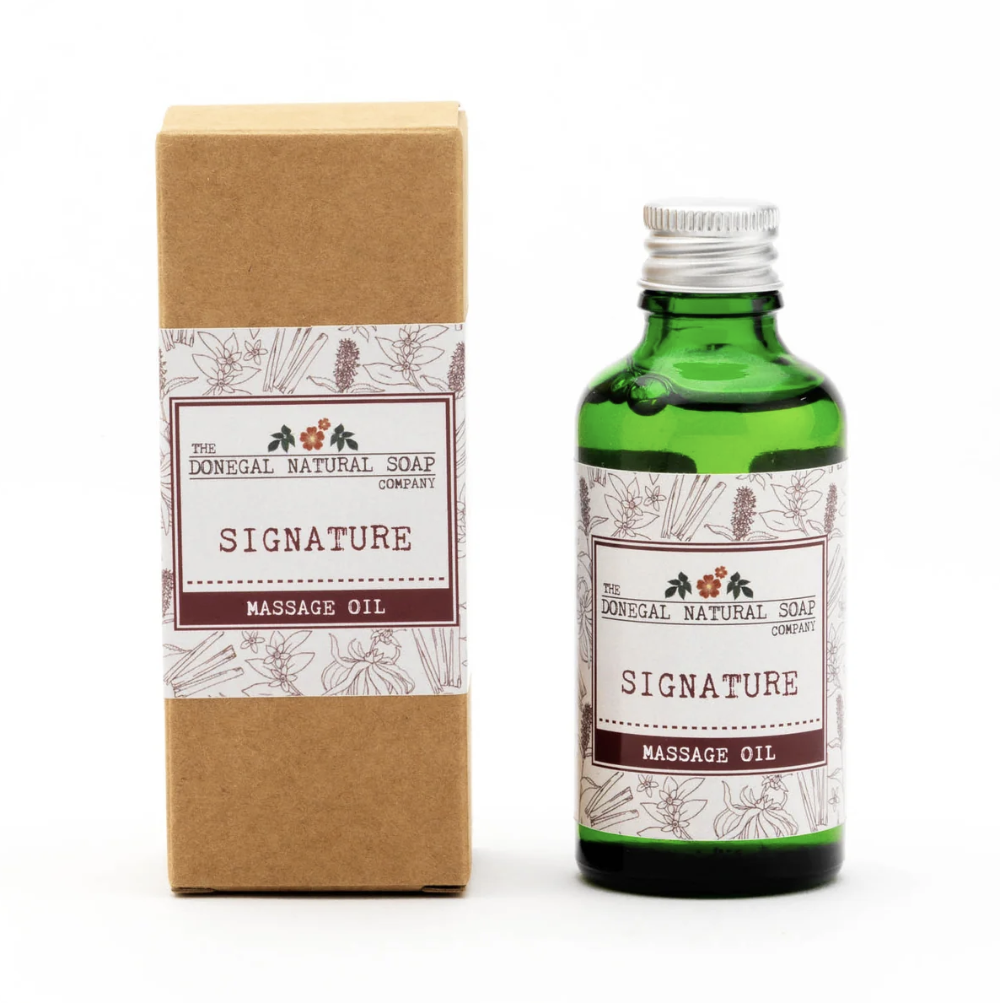 A green glass bottle of massage oil next to its brown kraft paper box packaging, both featuring a white label with botanical illustrations and the brand name 'The Donegal Natural Soap Company'.