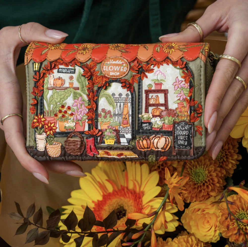 A person holds a small zip-around wallet decorated with an embroidered autumn flower shop scene. The wallet features orange pumpkins, fall leaves, flowers, and storefront details, with the words 'Flower Shop' visible in the embroidery.