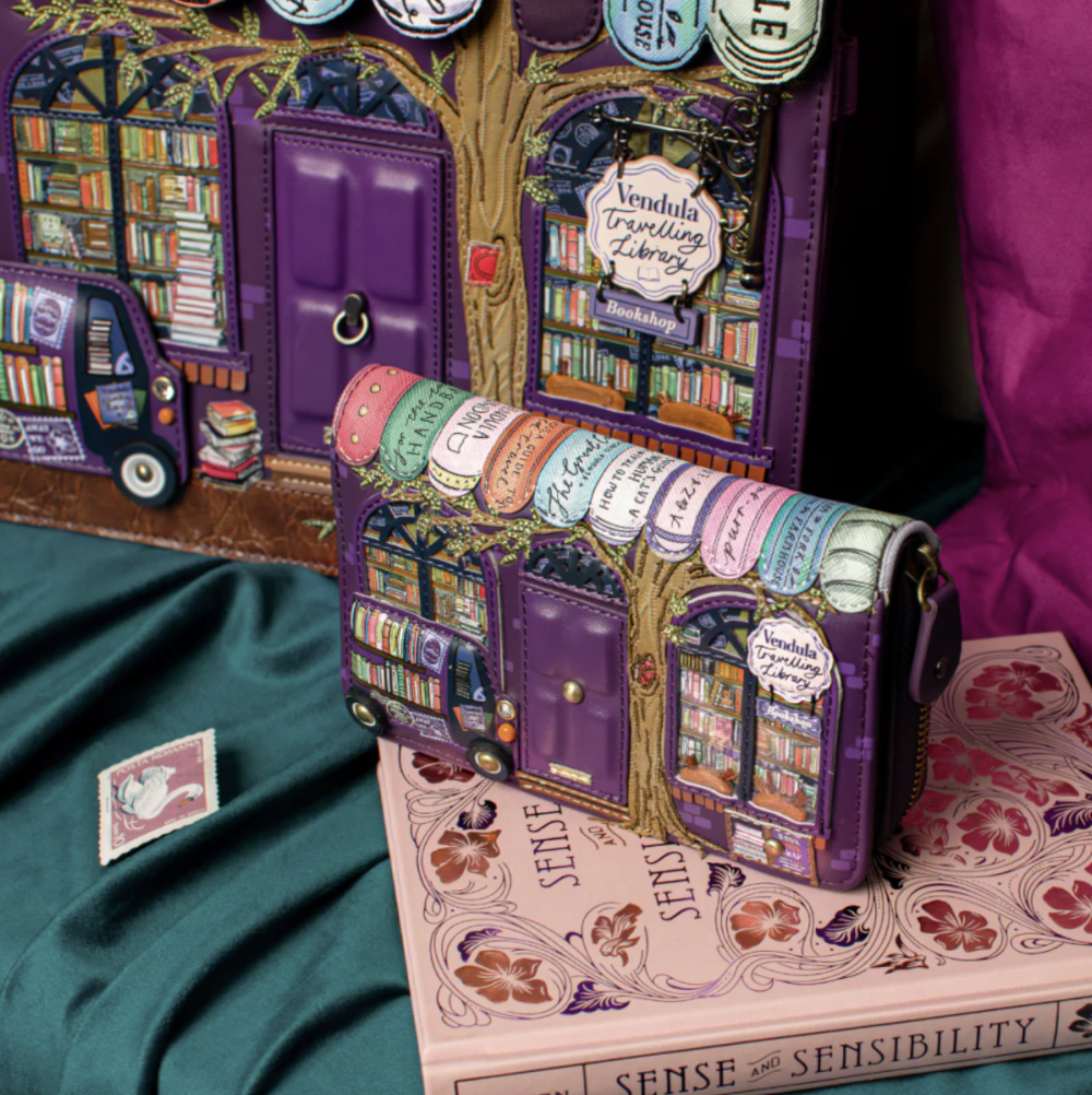 A purple and multicolor book bag displayed on top of a pink book, with a matching book-themed wallet behind it and a small matching pin nearby on a dark green satin surface.